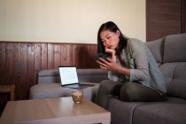 Young ethnic female with glass of coffee surfing internet on cellphone sitting in couch at home room near laptop — Stock Photo