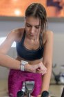 Concentrated sweaty young sportswoman in top and legging doing cardio training on exercise bike in gym checking smart watch — Stock Photo