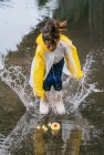 Charmant enfant ethnique en imperméable jouant avec des canards en plastique se reflétant dans la flaque ondulée par temps pluvieux — Photo de stock
