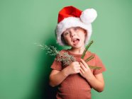 Garçon joyeux en chapeau de Père Noël rouge avec branches de sapin regardant la caméra avec les yeux grands ouverts — Photo de stock
