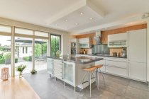 Cucina moderna con tavolo e sgabelli contro frigorifero e armadi in casa con parete di vetro durante il giorno — Foto stock