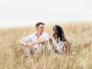 Homme souriant adulte avec une femme ethnique bien-aimée se reposant sur le pré tout en passant le week-end à la campagne — Photo de stock