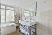 Біла ванна, розміщена біля вікна в стильній ванній кімнаті з чистими раковинами під дзеркалом і бежевими плитковими стінами в сучасній квартирі вдень — стокове фото