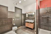 Combined bathroom with bathtub shower cabin and sink with double faucets with toilet near gray door under glowing lights — Stock Photo