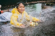 Charmantes ethnisches Kind im Regenmantel, das mit Plastikenten spielt, die bei Regenwetter in einer Pfütze reflektieren — Stockfoto