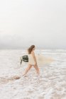 Side view of young sportswoman with flying hair and surfboard in ocean with foam under cloudy sky — Stock Photo
