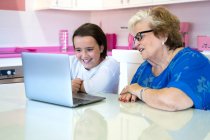 Smiling granddaughter and grandmother sitting at table and using laptop in light room in apartment — Stock Photo