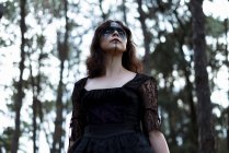Снизу мистическая ведьма в длинном черном платье и с раскрашенным лицом, смотрящим в темные мрачные леса — стоковое фото