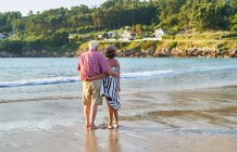 Back view of unrecognizable barefoot elderly couple in sunglasses standing on wet sandy beach and enjoying sunny day — Stock Photo