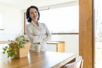 Mature confident woman standing looking at camera in kitchen near countertop holding mug of hot beverage in the morning at home — Stock Photo