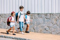 Schoolboy with backpack speaking with female friends while strolling on tiled pavement against stone wall in sunlight — Stock Photo
