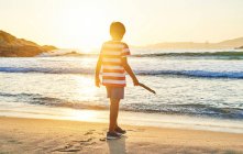 Вид ззаду на повну довжину невпізнаваного хлопчика з палицею, що стоїть на мокрому піщаному березі, промитому махаючи блакитним морем на заході сонця — стокове фото