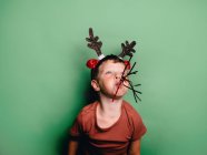 Menino vestindo chifres de rena headband e festa festiva ventilador na boca de pé contra o fundo verde e olhando para longe — Fotografia de Stock