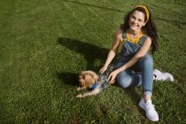 Высокий угол полного тела счастливой молодой женщины, сидящей на газоне со скрещенными ногами и гладящей йоркширского терьера, глядя в камеру — стоковое фото