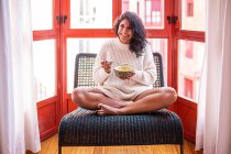 Pieno corpo di donna latina scalza seduta con le gambe incrociate distogliendo lo sguardo sulla sedia e mangiando zuppa dalla ciotola — Foto stock