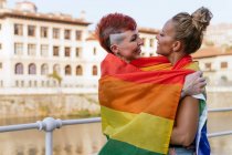 Cool mujer tatuada con mohawk y bandera LGBTQ abrazando novia con los ojos cerrados contra el canal en la ciudad - foto de stock