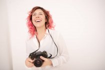 Sorria fotógrafa feminina com cabelo rosa segurando uma câmera fotográfica profissional em suas mãos na sala de luz — Fotografia de Stock