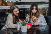 Las mejores amigas con vasos de bebidas refrescantes navegando en el teléfono celular en la mesa en la cafetería urbana - foto de stock