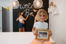 Charmantes Kind mit zukünftigem Geschwister-Ultraschallbild vor Kamera gegen schwangere Mutter und Vater, die auf Kreidetafel im Haus schreiben — Stockfoto