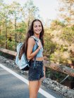 Side view of happy female traveler with rucksack looking at camera while walking on asphalt roadway in Tenerife Canary Islands Spain — Stock Photo