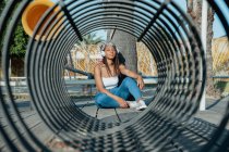 Fêmea étnica legal ouvindo música de fone de ouvido enquanto sentado com pernas cruzadas contra a construção em forma de túnel e olhando para a câmera — Fotografia de Stock