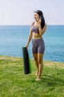 Sincere young ethnic female in sportswear with yoga mat roll speaking on cellphone while looking down on ocean beach — Stock Photo