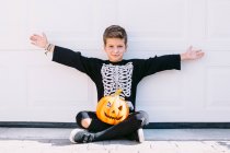 Cuerpo completo de niño excitado en traje de esqueleto con maquillaje y calabaza de Halloween tallada levantando brazos y gritando con cara de miedo mientras está sentado cerca de la pared blanca - foto de stock