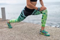 Recorte irreconocible corredor femenino estirando las piernas y haciendo ejercicio de embestida hacia adelante mientras se entrena en el paseo marítimo en verano - foto de stock