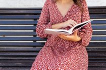 Обрезанная неузнаваемая женщина в стильной одежде сидит с открытой книгой на деревянной скамейке напротив здания со светлой стеной в дневное время — стоковое фото