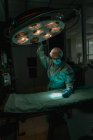 Joven veterinaria en uniforme quirúrgico y máscara estéril mirando hacia abajo mientras ajusta la lámpara por encima del textil médico en la clínica - foto de stock