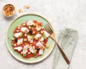 Tomate de verão e salada de bacalhau em um prato raso com um garfo e uma tigela de cebolas fritas — Fotografia de Stock