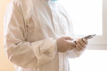Обрізаний невизначений чоловічий медик у особистому захисному обладнанні, який торкається екрана на смартфоні під час пандемії COVID 19 у клініці — стокове фото