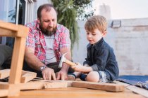 Barbudo pai ensinando filho com martelo trabalhando com madeira enquanto sentado no calçadão no fim de semana — Fotografia de Stock