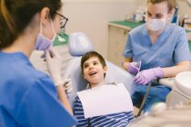 Alto ángulo de niño con la boca abierta hablando con los médicos durante el tratamiento dental en el consultorio dental contemporáneo - foto de stock