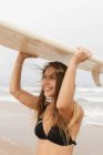 Jeune athlète souriante en maillot de bain avec des cheveux volants portant une planche de surf sur la tête regardant vers l'avant sur la côte océanique — Photo de stock
