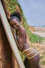 Seitenansicht des Inhalts junge afroamerikanische Surferin in Badebekleidung lehnt an Longboard-Surfen, während sie an der Küste in die Kamera schaut — Stockfoto