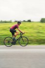 Motion blur side view of sportsman in protective helmet riding bicycle during workout on asphalt roadway against green hill and trees under light sky — Stock Photo