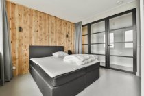 Cama confortável com linho branco colocado contra a parede de madeira no quarto com design minimalista — Fotografia de Stock