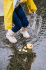 From above crop anonymous child in rubber boots having fun in puddle with splattering aqua on rainy day — Stock Photo