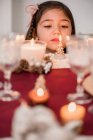 Нежный ребенок, созерцающий горящую свечу в стакане на столе с хвойными конусами во время новогоднего праздника дома — стоковое фото