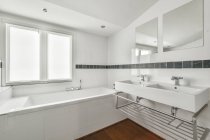 Lavabo double blanc sous des miroirs placés près de la baignoire dans une salle de bain spacieuse avec des murs carrelés blancs en journée — Photo de stock