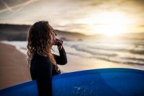 Vista lateral da jovem mulher de pé olhando para longe e assobiando na costa com prancha de surf antes de entrar no mar durante o pôr do sol na praia em Astúrias, Espanha — Fotografia de Stock