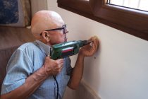 Concentrated old male in eyewear with electric screwdriver screwing plastic piece to wall in house — Stock Photo