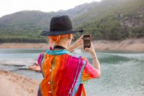 Visão traseira mulher com chapéu preto em um lago tirando fotos da paisagem com um celular — Fotografia de Stock
