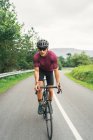 Desportista no capacete de proteção andar de bicicleta durante o treino na estrada de asfalto contra a colina verde e árvores sob o céu claro — Fotografia de Stock