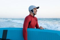 Vue latérale du surfeur masculin en combinaison et chapeau portant une planche à pagaie et entrant dans l'eau pour surfer sur le bord de la mer — Photo de stock