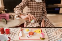 Crop bambino irriconoscibile in abito a scacchi con coltello giocattolo tagliere formaggio sul tagliere durante il gioco in casa — Foto stock