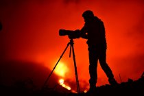 Vista lateral de la silueta de un hombre grabando y fotografiando con un trípode la explosión de lava en La Palma Islas Canarias 2021 - foto de stock