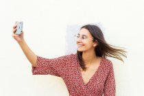 Mujer sonriente en vestido y gafas de pie tomando selfie con teléfono inteligente cerca de la pared blanca en el día - foto de stock