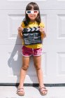 Menina feliz bonito em roupas coloridas casuais segurando clapperboard enquanto em óculos tridimensionais de pé no fundo da parede branca — Fotografia de Stock